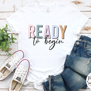 K&ouml;nnte beinhalten: Wei&szlig;es T-Shirt mit dem Text "READY to begin" in bunten Blockbuchstaben. Das Shirt liegt auf einer wei&szlig;en Holzoberfl&auml;che mit einem Paar wei&szlig;en Sneakers und einer blauen Jeans.