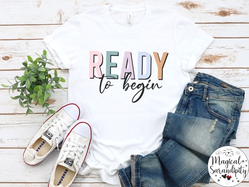 K&ouml;nnte beinhalten: Wei&szlig;es T-Shirt mit dem Text "READY to begin" in bunten Blockbuchstaben. Das Shirt liegt auf einer wei&szlig;en Holzoberfl&auml;che mit einem Paar wei&szlig;en Sneakers und einer blauen Jeans.