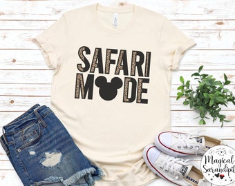 May include: A beige t-shirt with the text "SAFARI MODE" in black with a leopard print pattern. A black Mickey Mouse head is in the middle of the text.