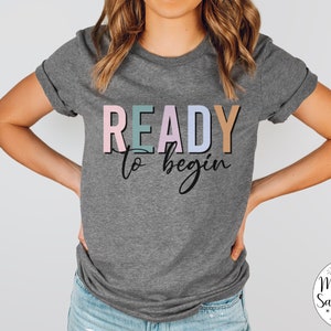 K&ouml;nnte beinhalten: Ein graues T-Shirt mit dem Text "READY to begin" in bunten Buchstaben. Die Buchstaben sind schwarz umrandet und haben einen leichten Schattensffekt. Der Text ist mittig auf dem Shirt platziert.