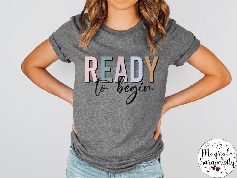 K&ouml;nnte beinhalten: Ein graues T-Shirt mit dem Text "READY to begin" in bunten Buchstaben. Die Buchstaben sind schwarz umrandet und haben einen leichten Schattensffekt. Der Text ist mittig auf dem Shirt platziert.