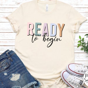 K&ouml;nnte beinhalten: Ein beiges T-Shirt mit dem Text "READY to begin" in bunten Blockbuchstaben. Die Buchstaben sind in den Farben Rosa, Blau, Gr&uuml;n und Orange.