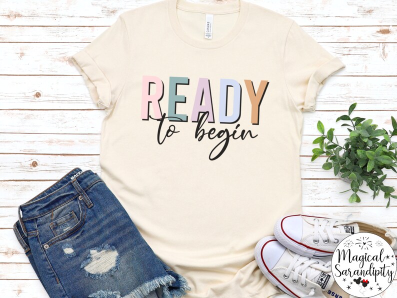K&ouml;nnte beinhalten: Ein beiges T-Shirt mit dem Text "READY to begin" in bunten Blockbuchstaben. Die Buchstaben sind in den Farben Rosa, Blau, Gr&uuml;n und Orange.