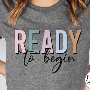 K&ouml;nnte beinhalten: Ein graues T-Shirt mit dem Text "READY to begin" in bunten Buchstaben. Der Text ist in einer verspielten Schriftart und die Farben sind pastellrosa, blau, gr&uuml;n und braun.