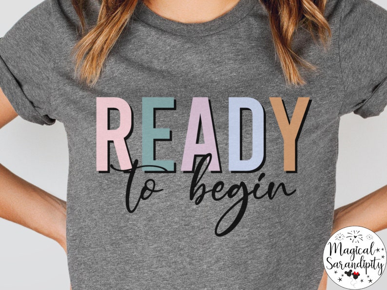 K&ouml;nnte beinhalten: Ein graues T-Shirt mit dem Text "READY to begin" in bunten Buchstaben. Der Text ist in einer verspielten Schriftart und die Farben sind pastellrosa, blau, gr&uuml;n und braun.