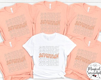 May include: Five t-shirts with the name "Savannah" printed in a retro font in a peach color and one white t-shirt with the same design. The name "Savannah" is printed in a peach color on the white t-shirt.