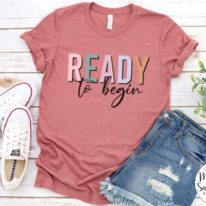 K&ouml;nnte beinhalten: Ein pinkfarbenes T-Shirt mit dem Text "READY to begin" in bunten Buchstaben. Das Shirt liegt auf einer wei&szlig;en Holzoberfl&auml;che mit einem Paar wei&szlig;en Sneakers und einer Jeans-Shorts.