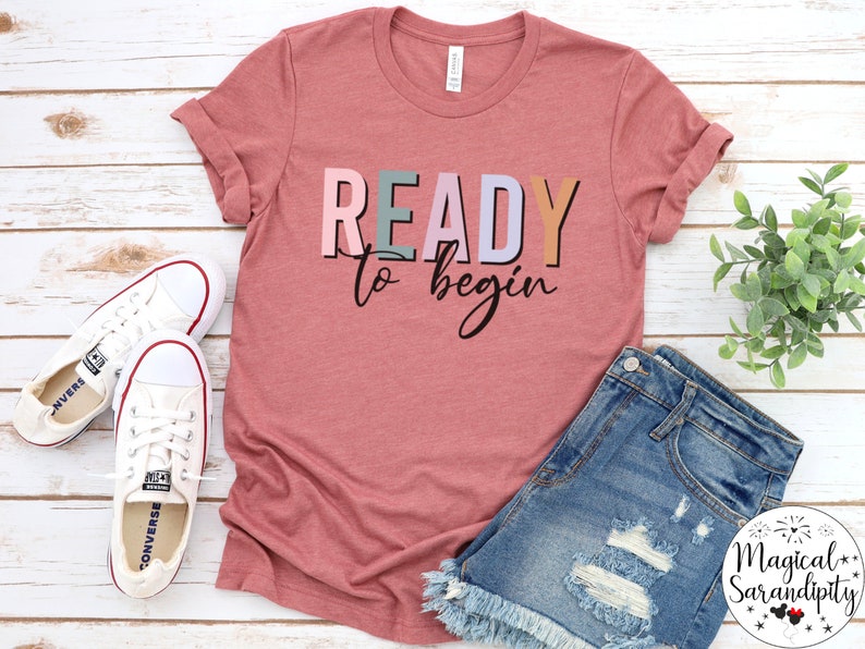 K&ouml;nnte beinhalten: Ein pinkfarbenes T-Shirt mit dem Text "READY to begin" in bunten Buchstaben. Das Shirt liegt auf einer wei&szlig;en Holzoberfl&auml;che mit einem Paar wei&szlig;en Sneakers und einer Jeans-Shorts.