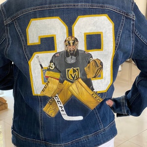 Op de afbeelding: Een blauwe denim jas met een wit en gouden ontwerp van een hockeykeeper met een helm en een hockeystick. Het nummer 29 is op de achterkant van de jas geschilderd.