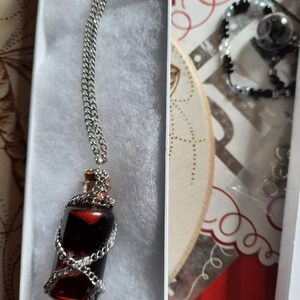 May include: A silver chain necklace with a red glass bottle pendant wrapped in silver wire. The bottle is filled with a dark red liquid.