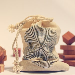 Pode incluir: Uma bolsa de lona branca com um padrão circular preto e branco. A bolsa é amarrada com um cordão branco e está sentada em uma mesa com blocos de madeira.