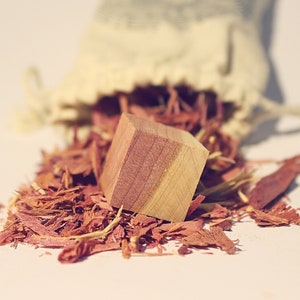 May include: A wooden cube with a reddish-brown hue sits on a bed of wood shavings. The shavings are a deep reddish-brown color and are scattered around the cube.