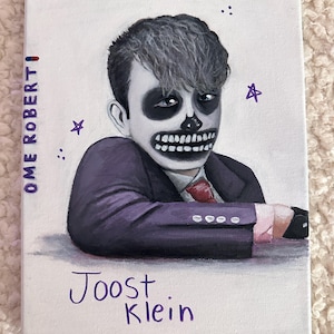 May include: A painting of a person with a skull face wearing a purple suit and a red tie. The painting is signed "Joost Klein" and has the words "Ome Robert" on the left side.