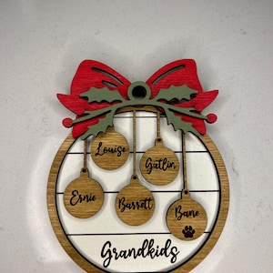 May include: A wooden Christmas ornament with a red bow and green holly leaves. The ornament has five hanging ornaments with the names "Louise", "Gatlin", "Ernie", "Barrett", and "Bane" written on them. The ornament also has the text "Grandkids" written on it.
