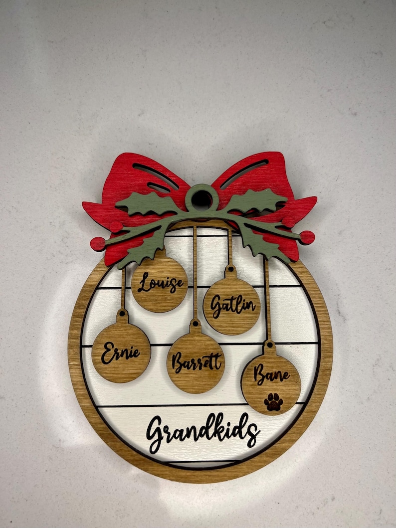 May include: A wooden Christmas ornament with a red bow and green holly leaves. The ornament has five hanging ornaments with the names "Louise", "Gatlin", "Ernie", "Barrett", and "Bane" written on them. The ornament also has the text "Grandkids" written on it.