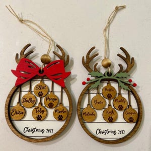 May include: Two wooden Christmas ornaments shaped like reindeer with a circle in the center. The circles have a white background with a wood grain pattern and are decorated with smaller circles that have names written on them. The ornaments are decorated with a red bow and green holly.