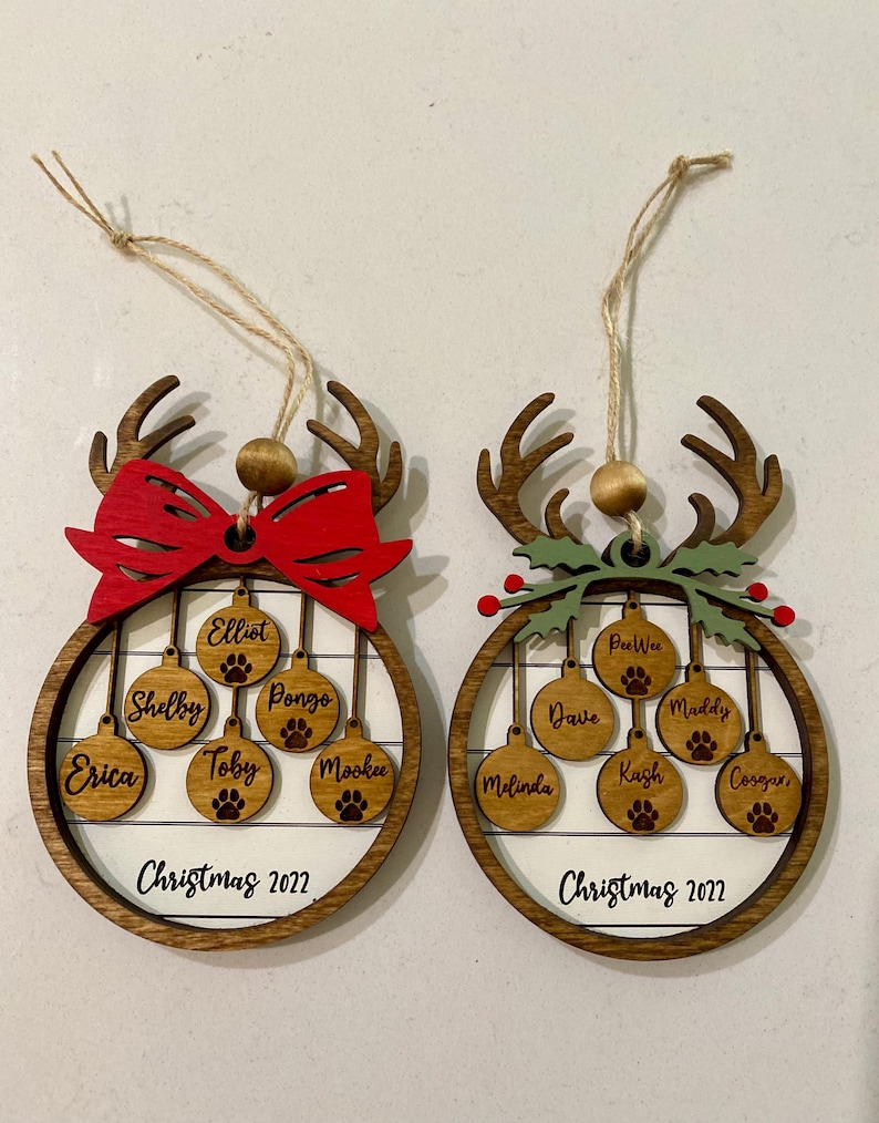 May include: Two wooden Christmas ornaments shaped like reindeer with a circle in the center. The circles have a white background with a wood grain pattern and are decorated with smaller circles that have names written on them. The ornaments are decorated with a red bow and green holly.