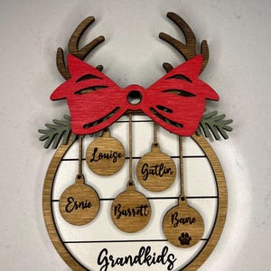 May include: A wooden Christmas ornament with a red bow and reindeer antlers. The ornament has a white background with the word "Grandkids" and five ornaments hanging from it with the names "Louise", "Gatlin", "Ernie", "Barrett", and "Bane".