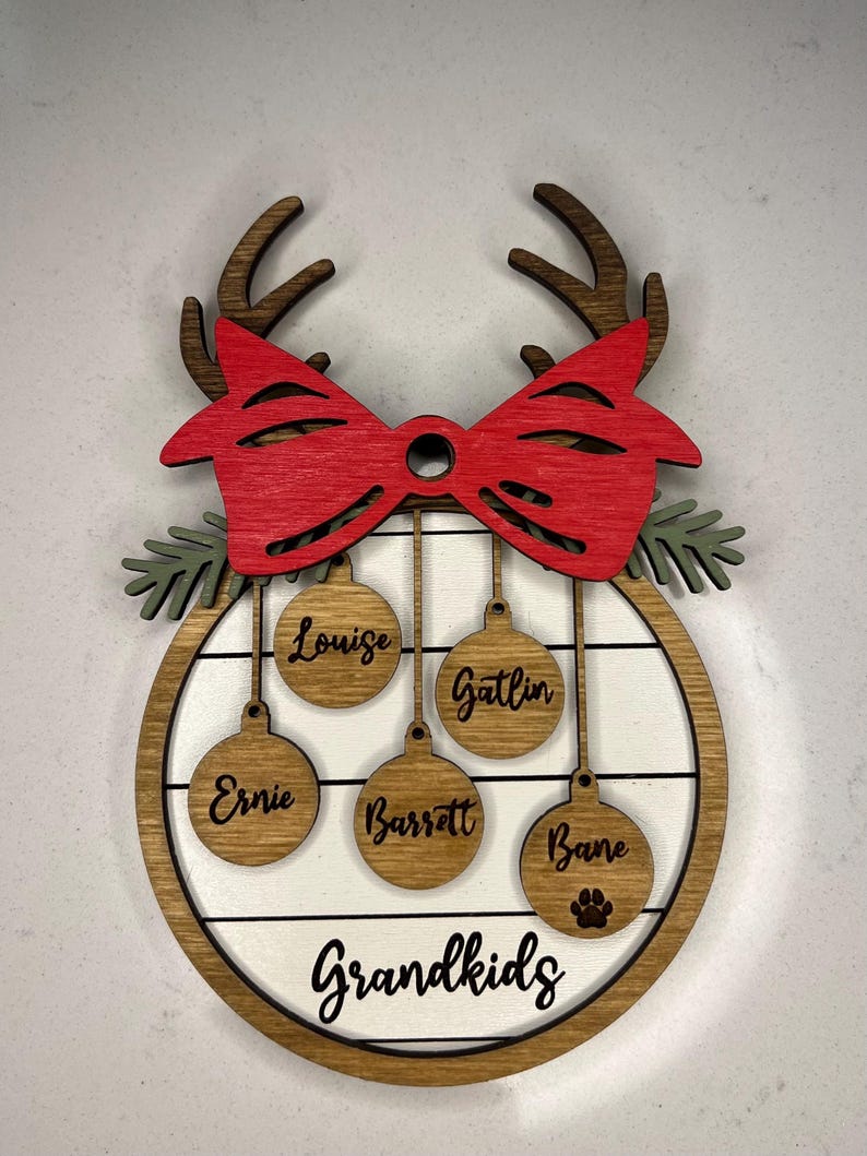 May include: A wooden Christmas ornament with a red bow and reindeer antlers. The ornament has a white background with the word "Grandkids" and five ornaments hanging from it with the names "Louise", "Gatlin", "Ernie", "Barrett", and "Bane".