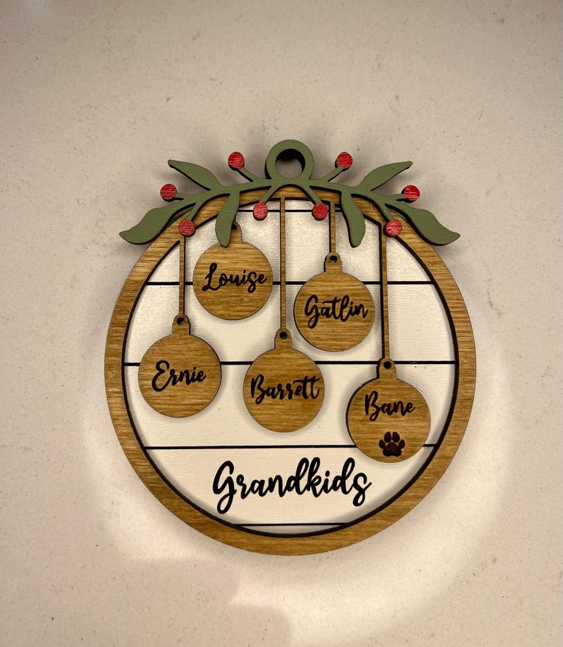 May include: A wooden Christmas ornament with a wreath and five ornaments hanging from it. The ornaments are personalized with the names "Louise", "Gatlin", "Ernie", "Barratt", and "Bane". The ornament also has the word "Grandkids" written on it.