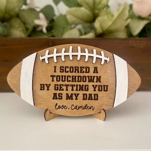 Puede incluir: Señal de madera con forma de balón de fútbol con el texto "Marqué un touchdown al tenerte como padre, con amor, Camden".