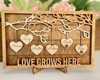 May include: A wooden sign with a carved tree branch and hanging hearts. The hearts are personalized with the names "Liz", "Aaron", "Austin", "Aiden", "Monte", and "Rex". The sign reads "LOVE GROWS HERE".