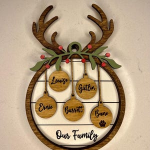 May include: Wooden Christmas ornament with reindeer antlers, a wreath, and five hanging ornaments with names "Louise", "Gatlin", "Ernie", "Barsett", and "Bane". The ornament has a white background with a woodgrain pattern and the text "Our Family" at the bottom.