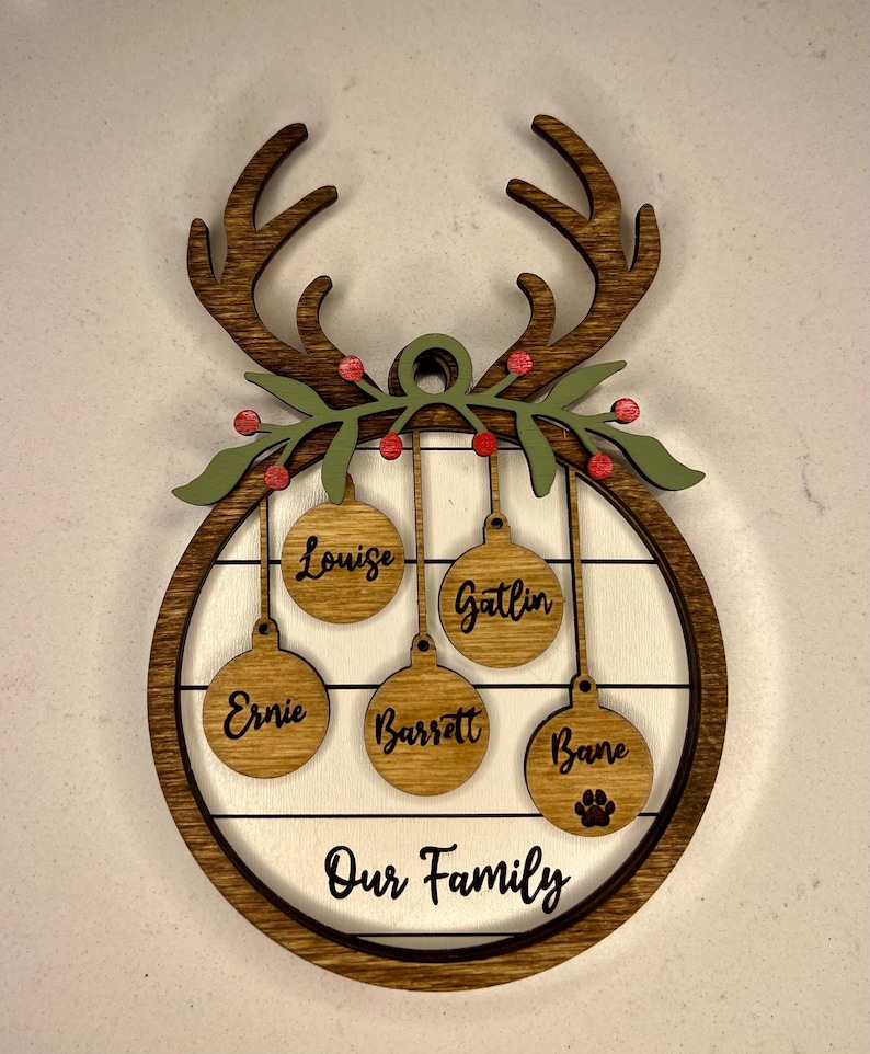 May include: Wooden Christmas ornament with reindeer antlers, a wreath, and five hanging ornaments with names "Louise", "Gatlin", "Ernie", "Barsett", and "Bane". The ornament has a white background with a woodgrain pattern and the text "Our Family" at the bottom.