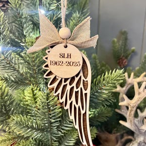 Può includere: Un ornamento natalizio a forma di ala d'angelo in legno chiaro, con fiocco beige e perla di legno. L'ornamento presenta le iniziali "SLH" e gli anni "1962-2025" incisi su un'etichetta circolare. È appeso a uno spago naturale.