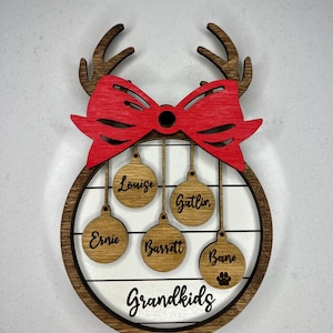 May include: A wooden Christmas ornament with a red bow and reindeer antlers. It has five round ornaments hanging from it with the names "Louise", "Gatlin", "Ernie", "Barrett", and "Bane" written on them. The bottom of the ornament says "Grandkids".