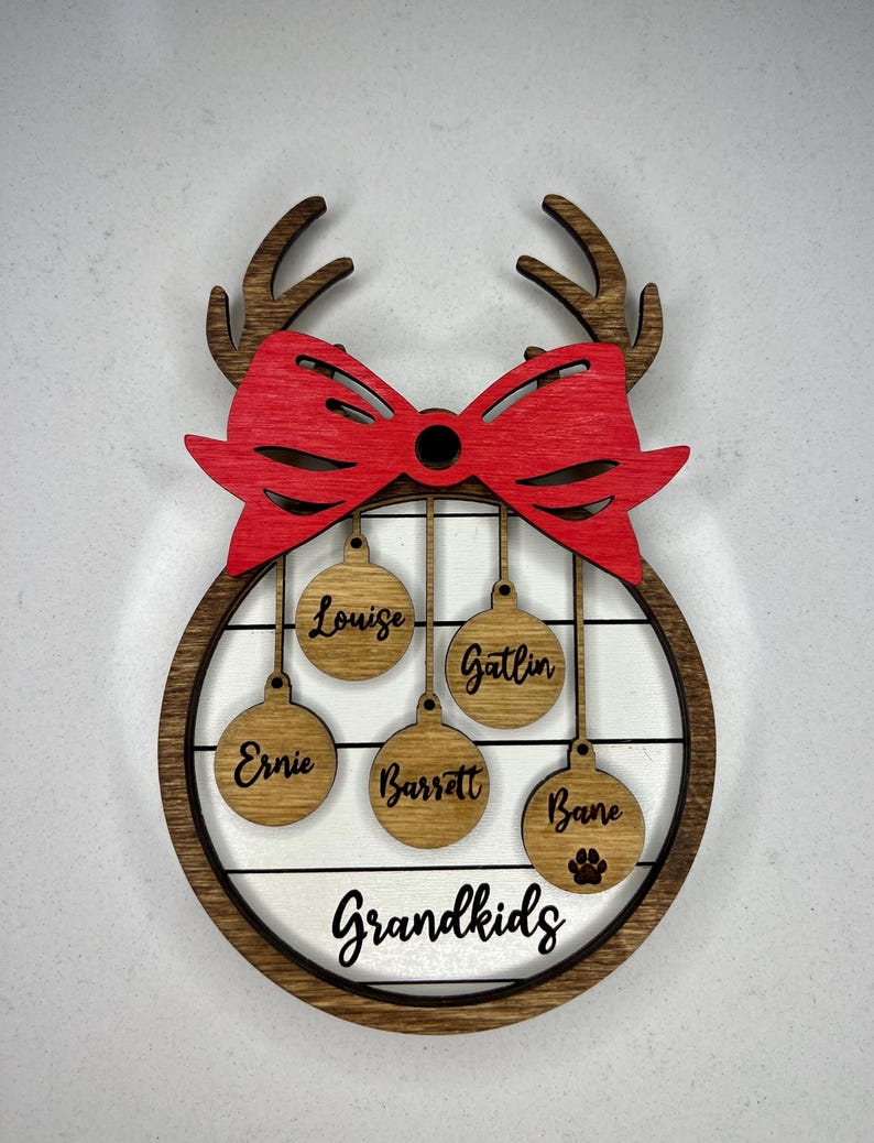 May include: A wooden Christmas ornament with a red bow and reindeer antlers. It has five round ornaments hanging from it with the names "Louise", "Gatlin", "Ernie", "Barrett", and "Bane" written on them. The bottom of the ornament says "Grandkids".