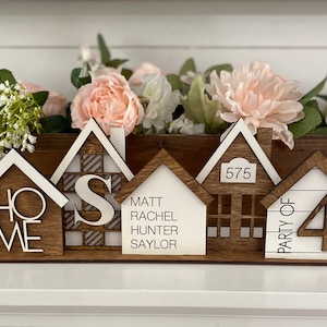 May include: Wooden house-shaped ornaments with the words "Home", "S", "Matt Rachel Hunter Saylor", "575", and "Party of 4" on them. The ornaments are arranged on a wooden base.