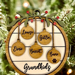 May include: A round wooden Christmas ornament with a white background and a green holly wreath at the top. The ornament has five hanging ornaments with names written on them: Louise, Gatlin, Ernie, Barrett, and Bane. The bottom of the ornament says "Grandkids".