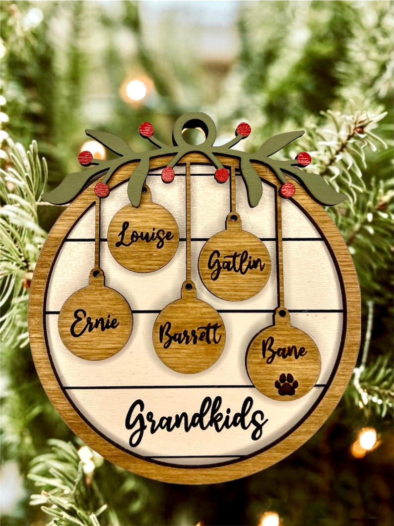 May include: A round wooden Christmas ornament with a white background and a green holly wreath at the top. The ornament has five hanging ornaments with names written on them: Louise, Gatlin, Ernie, Barrett, and Bane. The bottom of the ornament says "Grandkids".