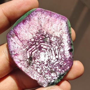 Puede incluir: Una rodaja pulida de piedra preciosa que presenta un centro vibrante en tonos púrpura y magenta con un patrón hexagonal de inclusiones blancas, rodeado por un borde exterior verde oscuro con una textura rugosa y natural.