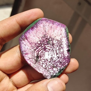 Pode incluir: Uma mão segura uma pedra preciosa polida de formato irregular. A pedra apresenta um interior roxo vibrante com veios brancos e uma borda verde. A superfície tem uma aparência texturizada, captando a luz.