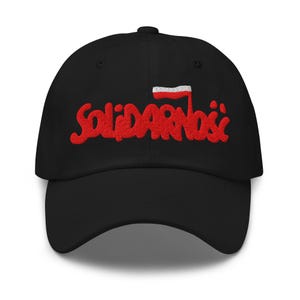 May include: Black baseball cap featuring the red embroidered word "Solidarność" and a small Polish flag. The cap has a curved peak and an adjustable strap. The design is a tribute to the Polish Solidarity movement.