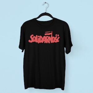 May include: Black t-shirt featuring the Polish word "Solidarność" in red, stylized lettering, with a red flag graphic. The shirt hangs on a black hanger against a light blue backdrop.