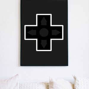 May include: Black framed wall art featuring a directional pad design. The artwork has a black background with a white-bordered cross shape. The cross contains arrows pointing up, down, left, and right, with a central circle.