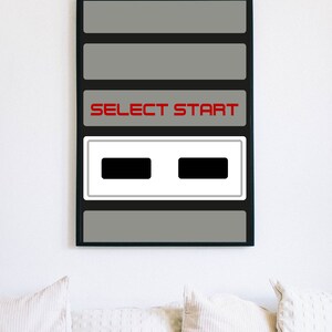 May include: Framed print featuring a retro video game controller design. The artwork displays a gray background with the words "SELECT START" in red. Below the text are two black rectangles. The print is displayed on a white wall.