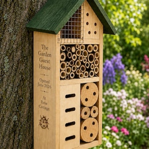 May include: A wooden insect hotel with a green roof, featuring a wire mesh section, bamboo tubes, a panel with horizontal slots, and circular wooden discs with holes. The side is engraved with The Garden Guest House, Opened June 2026, At Rose Cottage.