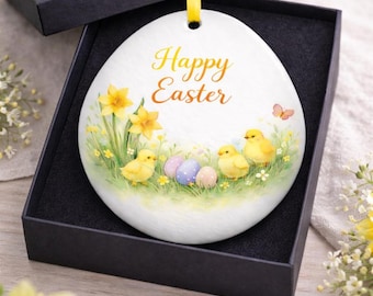 May include: A white, egg-shaped ornament with the words "Happy Easter" in yellow script. The ornament features a spring scene with yellow chicks, daffodils, and pastel Easter eggs. It is presented in a black gift box with a yellow ribbon.