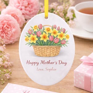 Pode incluir: Enfeite oval de cerâmica com uma fita amarela. O enfeite apresenta uma ilustração em aquarela de uma cesta cheia de narcisos amarelos e tulipas cor-de-rosa. O texto diz "Happy Mother's Day, Love, Sophie."