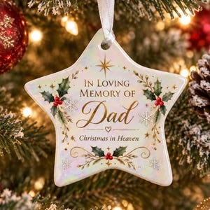 May include: A star-shaped ceramic ornament with an iridescent finish, featuring the text "In Loving Memory of Dad" and "Christmas in Heaven" in gold lettering, decorated with green holly leaves, red berries, and small white snowflake accents.