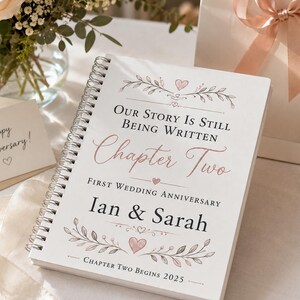May include: A white spiral-bound notebook with a rose gold and black floral design. The cover text reads: Our Story Is Still Being Written, Chapter Two, First Wedding Anniversary, Ian & Sarah, Chapter Two Begins 2025, decorated with small hearts.