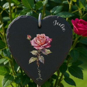 May include: A rose bush in a pot, displaying bright pink flowers and green foliage. A heart-shaped slate garden marker, featuring a rose illustration and the names "Julie" and "June", is placed in front. The pot is a light beige colour.