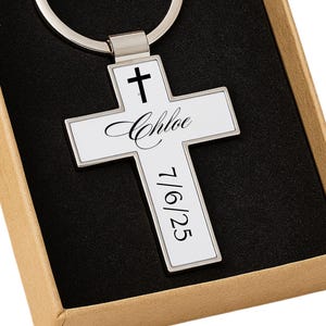 May include: A silver cross-shaped keyring with the name "Chloe" and the date "7/6/25" printed on it. The keyring is presented in a brown gift box with a black interior. The cross features a small cross symbol at the top.