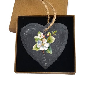 Puede incluir: Una placa de pizarra negra en forma de corazón con una imagen pintada de flores de manzano blancas y hojas verdes. La placa tiene el texto "Gracias" y "Sra. Smith" escrito en ella. La placa está en una caja marrón con un forro de terciopelo negro.