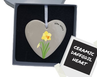 May include: A heart-shaped ceramic ornament in a gray gift box. The ornament features a hand-painted yellow daffodil with green leaves and the text "Ellie 2021". A polaroid-style card reads "CERAMIC DAFFODIL HEART".