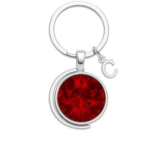 May include: A silver keyring featuring a round, faceted, deep red pendant. A small silver letter "C" charm hangs from the keyring. The pendant has a shiny, reflective surface.